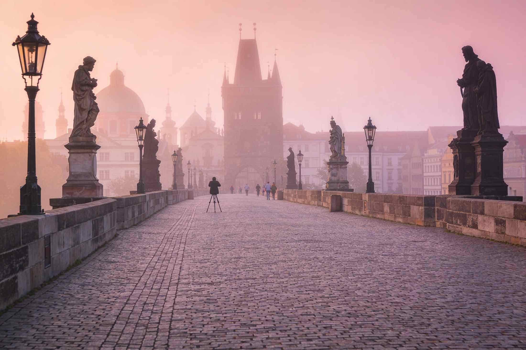 Charles Bridge almost empty at dawn in soft pink mist, a photographer with a tripod far along it