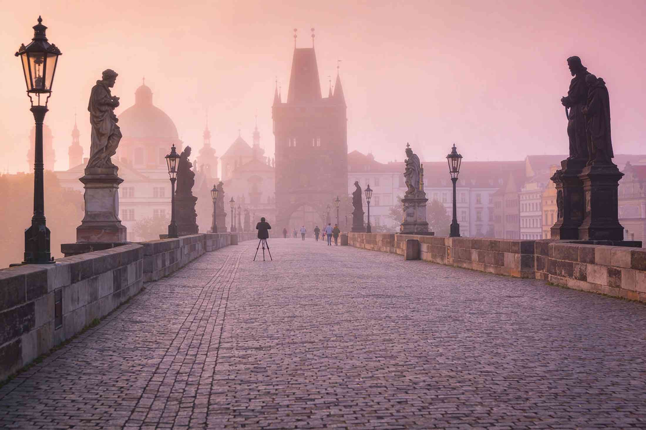 Charles Bridge almost empty at dawn in soft pink mist, a photographer with a tripod far along it