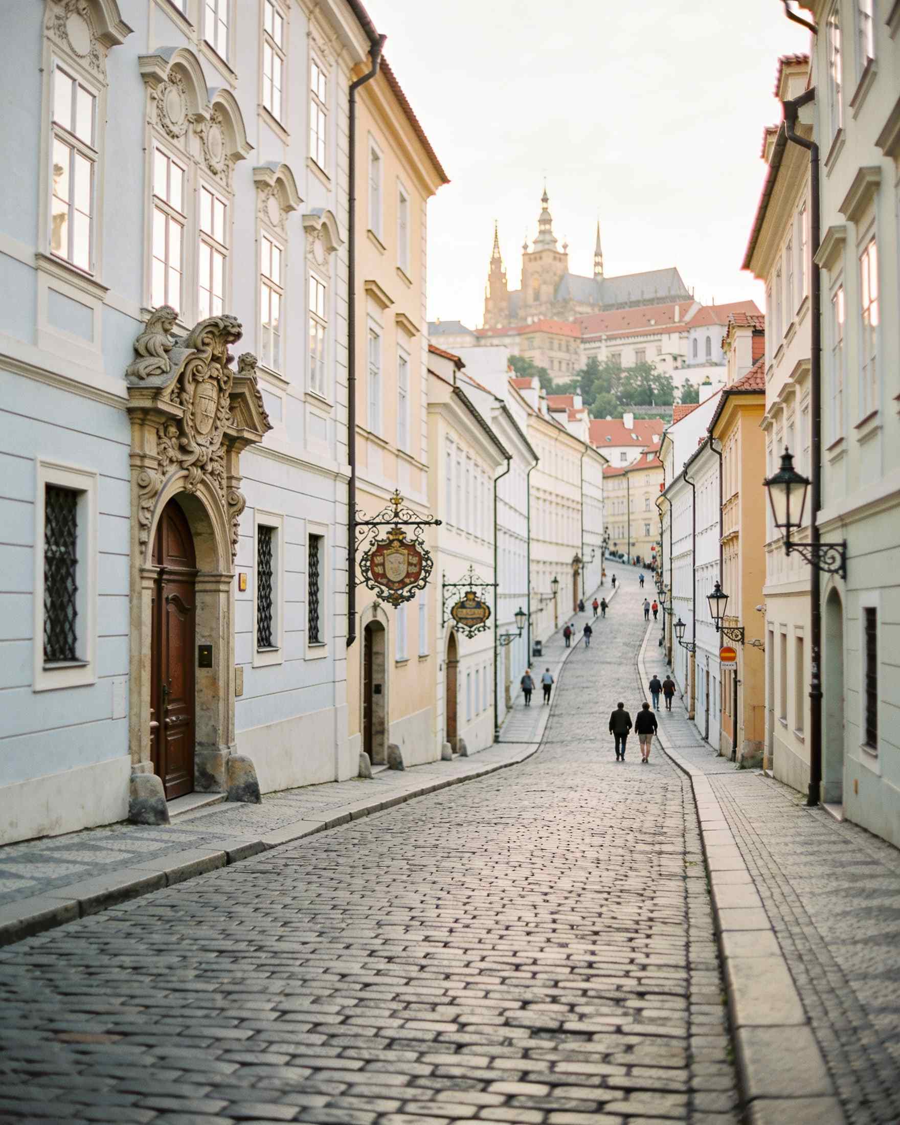 Nerudova Street climbing towards the castle between pastel baroque houses with carved house signs