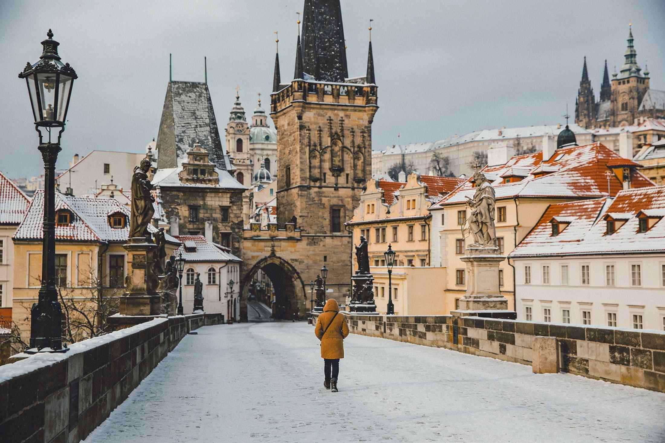 A walker in a warm coat on snowy Charles Bridge, the bridge tower and snowy roofs ahead
