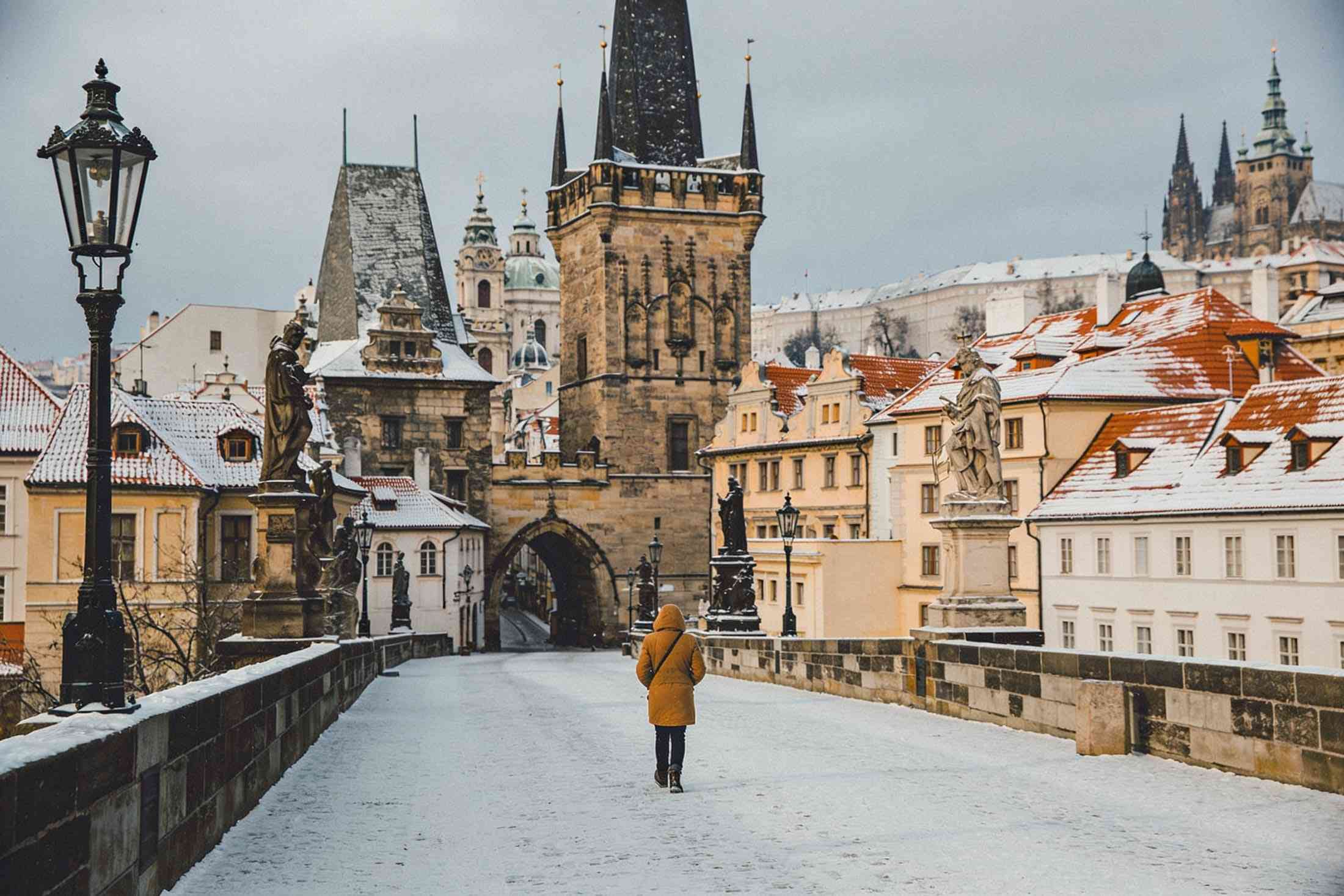 A walker in a warm coat on snowy Charles Bridge, the bridge tower and snowy roofs ahead